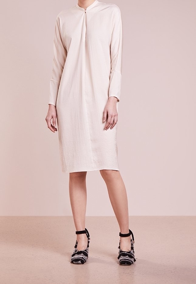Femme portant une robe beige clair longueur genoux à manches longues avec col montant, associée à des talons texturés noirs et blancs à bride à la cheville.