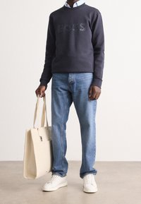 Marintröja med präglad "BOSS"-logotyp, ljusblå jeans, vita sneakers och en krämfärgad tote-väska med handtag. Slät textur, avslappnad design.