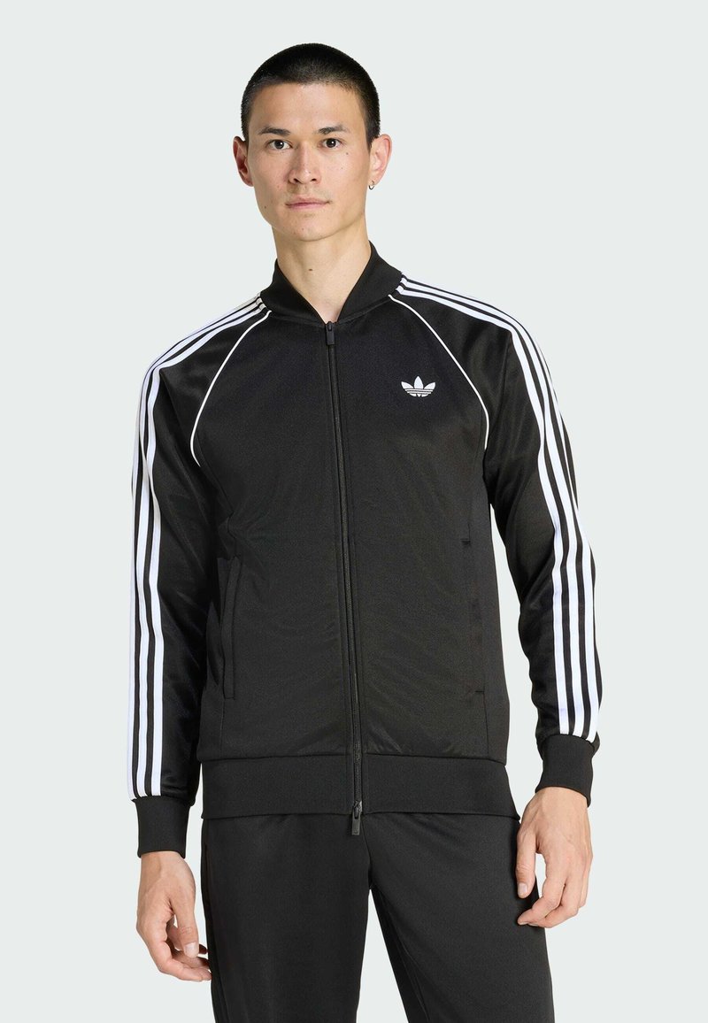 Άνδρας που φορά μαύρη ζακέτα σπορ Adidas με λευκές ρίγες κατά μήκος των μανικιών και το λογότυπο της Adidas στο στήθος, στέκεται μπροστά από απλό φόντο.