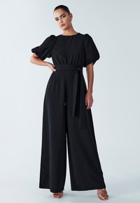 Zwarte jumpsuit met pofmouwen, getailleerde taille met een strik, wijd uitlopende pijpen, gladde textuur en enkelhoge openingen gecombineerd met sandalen met bandjes.