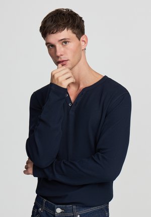 Maglietta henley a maniche lunghe di colore blu navy in tessuto waffle, con scollatura a V e patta di bottoni, abbinata a jeans in denim scuro.