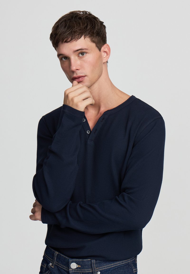 Maglietta henley a maniche lunghe di colore blu navy in tessuto waffle, con scollatura a V e patta di bottoni, abbinata a jeans in denim scuro.