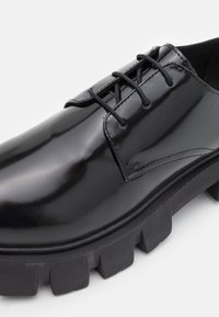 Černá lakovaná kožená bota s kulatou špičkou, šněrovacím designem a silnou, texturovanou gumovou podešví. Obsahuje minimální detaily šití.