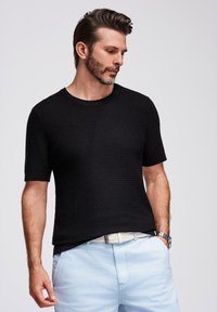 Maglione corto a maniche corte di colore nero, con un collo a giro, abbinato a pantaloni corti azzurri e una cintura decorativa.