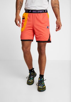 Mężczyzna ubrany w jaskrawo koralowe i żółte sportowe spodenki Nike z czarnym wykończeniem oraz wielokolorowe buty sportowe, stojący na jednolitym tle.