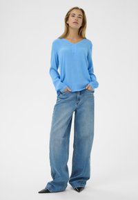 Haut bleu clair à manches longues avec un col en V et une patte de boutonnage discrète à l'avant, associé à un jean denim à jambes larges et des chaussures noires à bouts pointus.