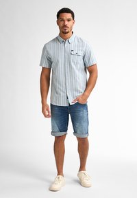 Lichtblauw overhemd met korte mouwen en verticale strepen, borstzak, gecombineerd met opgerolde denim shorts en beige sneakers. Casual zomeroutfit.