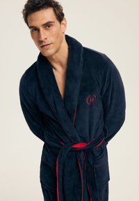 Młody mężczyzna w ciemnoniebieskim, pluszowym szlafroku z czerwonym wykończeniem, związanym w talii, stojący na jednolitym jasnym tle.