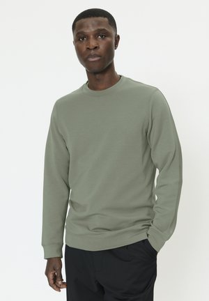 Muž oblečený v svetlo olivovozelenej dlhoprstej mikine s čiernymi nohavicami stojaci pred jednoduchým bielym pozadím.