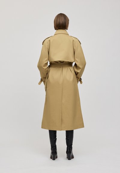 Cappotto beige con vestibilità su misura, vita cintrata e spalline sulle spalle. La texture liscia e il design a lunghezza media mettono in risalto linee nette.