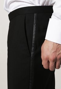 Homme portant un pantalon de costume noir avec une bande satinée sur le côté et une chemise blanche avec le poignet visible près de la main.