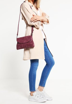 Mujer de pie con los brazos cruzados, vistiendo un abrigo beige tipo trench, jeans ajustados azules, zapatillas blancas y un bolso cruzado morado.