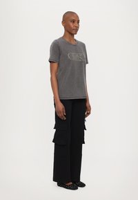 Camiseta de algodón gris con mangas cortas y cuello redondo, que presenta un diseño de logotipo texturizado. Combinada con pantalones cargo negros y zapatos planos.