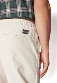Shorts beiges avec une ceinture élastique, comportant une poche arrière et une étiquette noire avec un texte blanc. Le tissu semble lisse et léger.