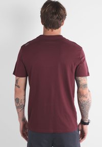 T-shirt en coton bordeaux avec un col rond, des manches courtes et un détail de design subtil à l'épaule. Texture lisse, coupe décontractée.