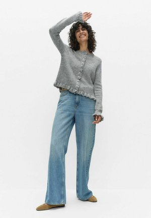 Grå stickad kofta med vågig fåll och knapprad, kombinerad med ljusblå jeans med vida ben och beige skor.