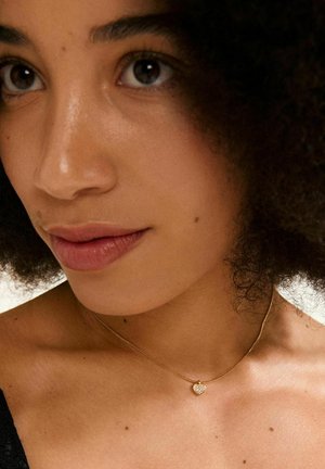 Collier en or avec un pendentif en forme de cœur, doté d'une surface texturée. Le pendentif est orné de petits détails scintillants, reposant sur une peau nue.