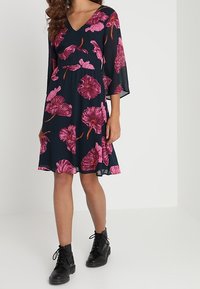 Marineblauwe jurk met roze bloemenprint en V-hals, met driekwart mouwen en een uitlopende rok. Gekleed met zwarte enkelboots.