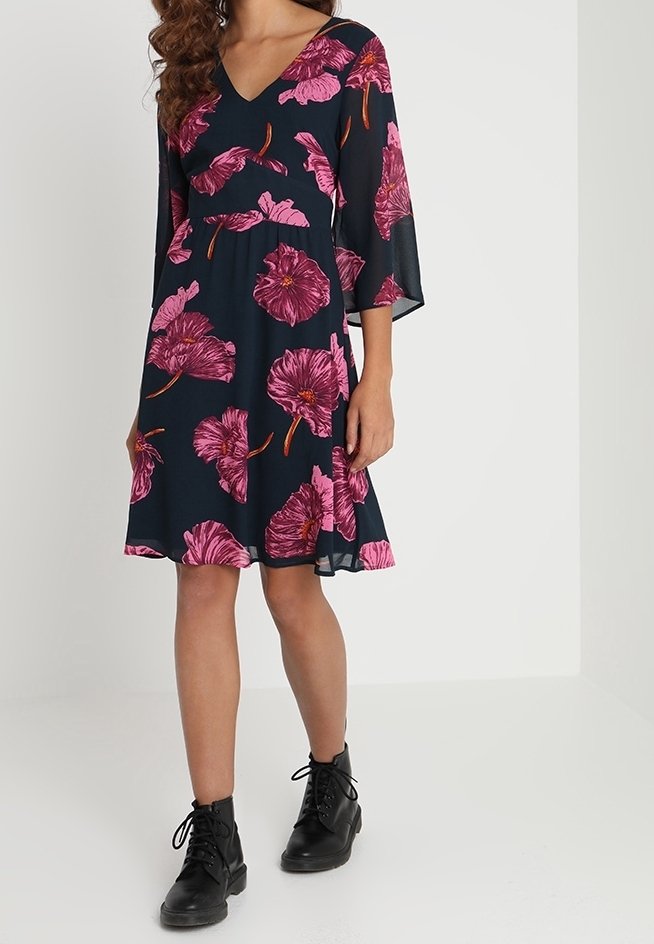 Marineblauwe jurk met roze bloemenprint en V-hals, met driekwart mouwen en een uitlopende rok. Gekleed met zwarte enkelboots.