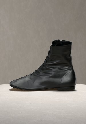 Bottine en cuir souple noir avec fermeture à boutons à l'avant, semelle plate et texture légèrement froissée, présentée sur une surface en pierre claire.