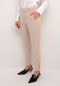 Beige kalhoty na míru s bočními kapsami a zipy na kotnících, spárované s černými nazouvacími botami s úzkým špičkovým tvarem. Hladká textilie.