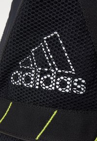 Svart nätlöparsko med texturerad yta, utrustad med en framträdande vit logotyp och en kontrasterande gul söm på den svarta remmen.