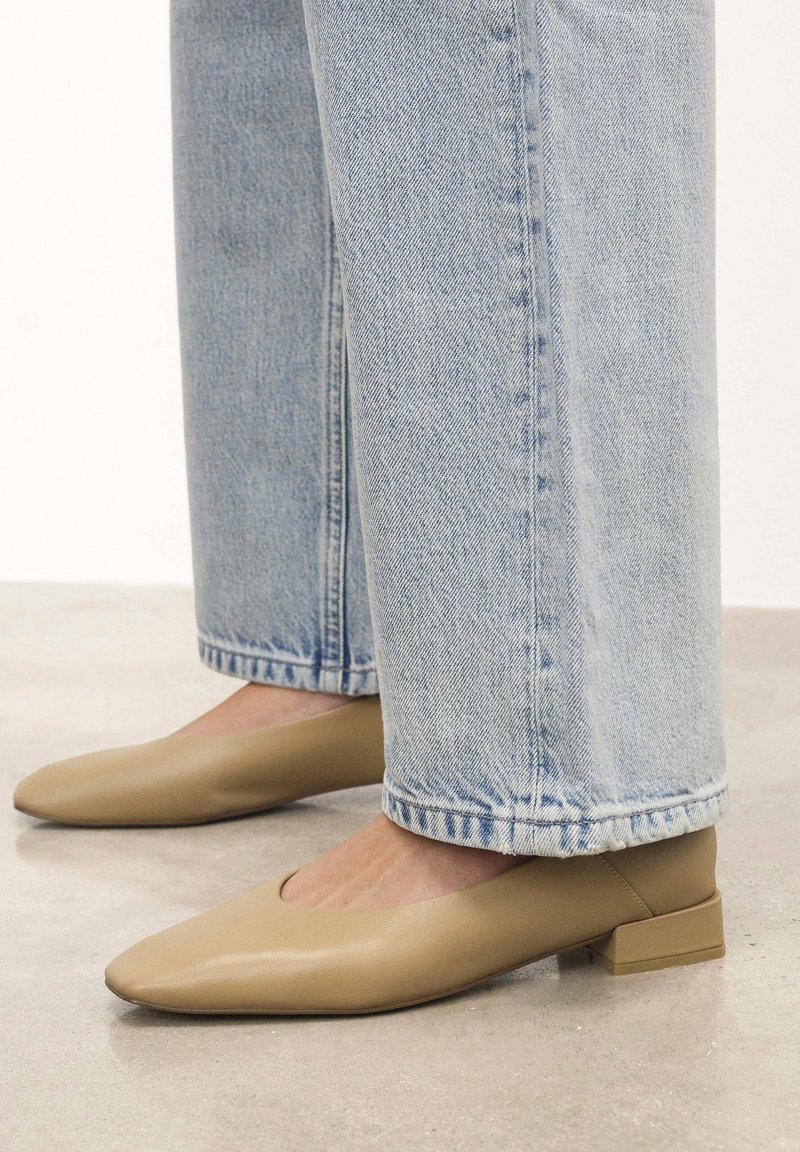 Bailarinas de cuero beige con punta afilada y un tacón cuadrado bajo, combinadas con unos vaqueros de pierna recta de color azul claro, sobre una superficie del suelo neutra.