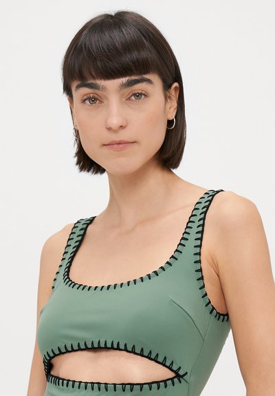 Zaļa peldkostīma topiņš ar melniem šuvēm, ar kvadrātveida izgriezumu un izgriezumiem sānos. Izgatavots no gludas, elastīgas vielas.