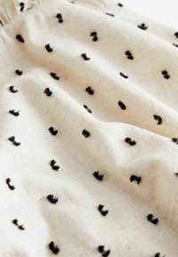 Tela de algodón en blanco roto con puntos dispersos de flecos negros. La textura está ligeramente arrugada, realzando su apariencia suave.