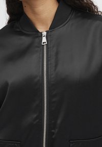 Schwarze Bomberjacke mit glattem Satin-Finish, geripptem Kragen und silbernem Metallreißverschluss sowie Taschen auf beiden Seiten.