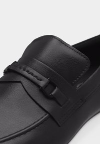 Mocassin en cuir noir avec une lanière décorative ornée d'une petite boucle rectangulaire en métal sur le devant supérieur.