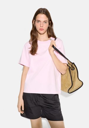 Młoda kobieta w jasnoróżowym T-shircie i czarnych szortach, nosi na ramieniu jasnobrązową zamszową torebkę z czarną lamówką.