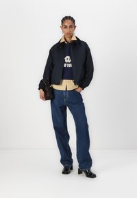 Navy bomber jacket over a navy knitted sweater, beige shirt, and loose blue denim jeans. Black heeled boots and a small brown bag.