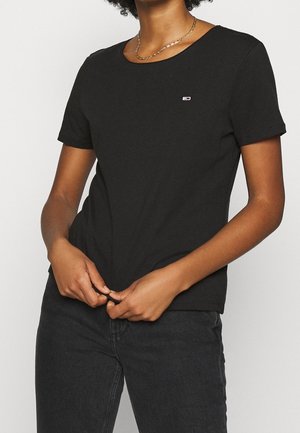 Persoon draagt een zwart T-shirt met korte mouwen en een klein logo op de borst, gecombineerd met een zwarte spijkerbroek en een gouden ketting.