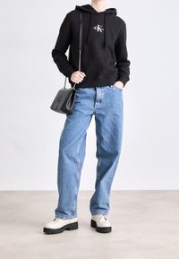 Felpa nera con cappuccio e logo, jeans blu chiaro a vestibilità rilassata e scarpe color crema con suole nere. Piccola borsa a tracolla nera con tracolla a catena.