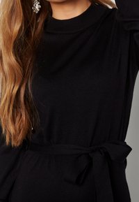 Pull noir avec un col montant et des manches longues, doté d'une ceinture à la taille. Texture douce, coupe ajustée et finition lisse sans motifs visibles.