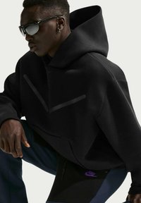 Μαύρο φαρδύ φούτερ με κουκούλα, με εντυπωσιακό V-σχήματος σχέδιο σε ματ φινίρισμα. Φορεμένο με να υτικά παντελόνια και μαύρα γυαλιά ηλίου. Φανερώνει τη σήμανση της Nike.