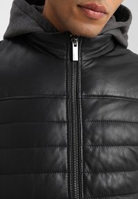 Man som bär en svart vadderad läderjacka med silverdragkedja och grå huvtröja under, närbild på bröstet och läpparna.