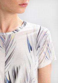 Halenka s krátkými rukávy vyrobená z lehkého, hladkého materiálu, zdobená abstraktním vzorem ve stylu akvarelu v šedé, modré a žluté barvě. Kulatý výstřih.