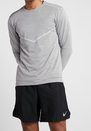 Mand iført en grå langærmet sportsbluse og sorte Nike-shorts med en grå swoosh-logo, stående med hænderne bag ryggen.