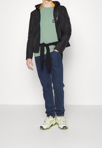 Svart lättviktsjacka med orange inredning, grön t-shirt, knuten svart tröja, jeans och kraftiga vita och gröna sneakers.