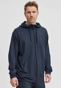 Námořnická mikina na zip vyrobená z hladkého materiálu, s klokaní kapsou, nastavitelnou kapucí se šňůrkou a minimalistickým logem na hrudi.