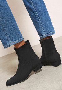 Melnas zamšādas potīšu zābaki ar dauzītu purngalu un blokveida papēdi. Sānu rāvējslēdzējs; apvienoti ar gaiši ziliem džinsiem.