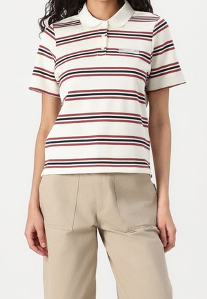 Vrouw draagt een wit poloshirt met rode en zwarte horizontale strepen en beige broek, staande met haar armen ontspannen langs haar zij.