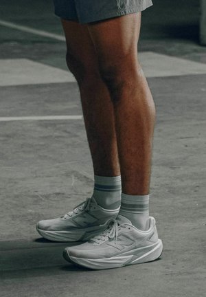 Beine, die graue Shorts, weiße Sportsocken und weiße New Balance Laufschuhe tragen, stehen auf einem grauen Betonboden.