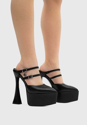 Scarpe platform nere con tacco largo a blocco e doppia fibbia, indossate a piedi nudi su sfondo semplice.