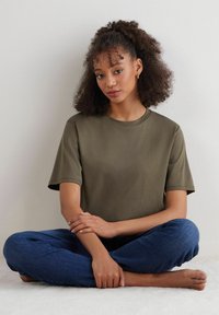 Olivgrünes Kurzarm-T-Shirt mit rundem Halsausschnitt, kombiniert mit blauen Jeans. Modell sitzt, die Hände auf den Knien ruhend, mit lockigem Haar.