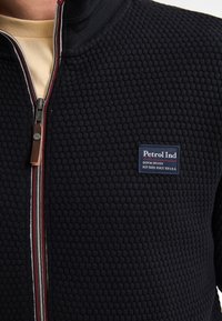 Giacca nera con texture, con colletto alto, dotata di zip frontale e tirazip in pelle. Un'etichetta blu è cucita sul petto.
