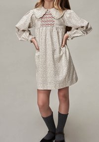 Sukienka w kwiatowy wzór z okrągłym kołnierzem, czerwonym marszczeniem na piersi, długimi bufkami i marszczoną talią; noszona z szarymi zakolanówkami.