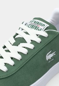 Grön mockaskosko med vita snören, med en Lacoste-logga på sidan och en strukturerad yta. Klassisk design med låg silhuett.